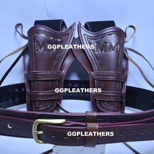 Może przedstawiać: Brązowy skórzany zestaw kabur na broń z dwiema kaburami i paskiem. Kabury są wytłoczone literami "MM" i nazwą marki "GGLEATHERS". Pasek ma złotą klamrę.