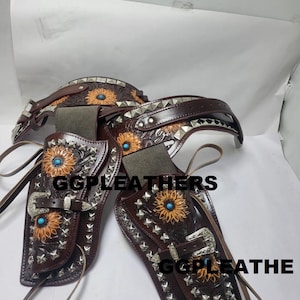 以下が含まれることがあります： 茶色の革製ホルスターセット。ひまわりとターコイズのアクセント、シルバースタッド、そして「GGPLEATHERS」の文字が特徴です。ベルトと2つのホルスターが含まれ、ウェスタンテーマを表現しています。