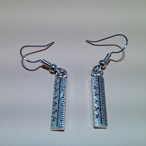 May include: A pair of silver-toned ruler earrings. Each earring features a small, rectangular ruler charm with etched markings and numbers. The earrings have a simple hook design for pierced ears. The background is a solid, neutral color.