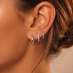 May include: Three silver hoop earrings are displayed on an ear. The first is a teardrop shape, the second is a small hoop with clear stones, and the third is a simple, small hoop. The earrings are shiny and reflect light.