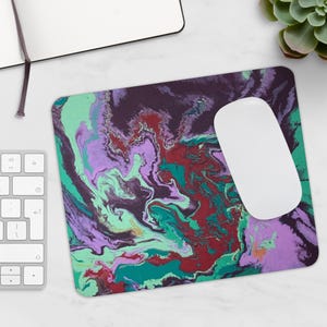 Peut inclure: Tapis de souris rectangulaire avec un motif abstrait tourbillonnant dans des tons de violet, vert et rouge. Une souris d'ordinateur blanche est posée dessus. Un clavier et un bloc-notes sont visibles.