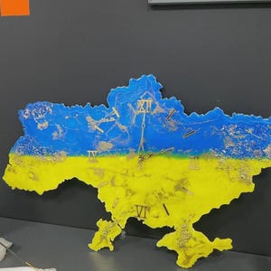 May include: A wall clock shaped like the map of Ukraine, featuring a blue and yellow color scheme. The clock has gold-colored Roman numeral hour markers and clock hands. The clock is mounted on a dark gray wall.