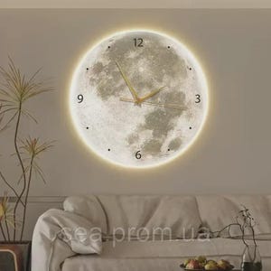 May include: A round wall clock with a moon design. The clock face is a detailed image of the moon, with a glowing halo around the edge. The clock hands are gold, and the numbers are indicated by dots and the numbers 3, 6, 9, and 12.