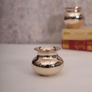 May include: A small, shiny, gold-colored pot with a wide rim and rounded body. The pot reflects the surrounding environment, creating a mirrored effect. Another similar pot is visible in the background, along with a stack of books.