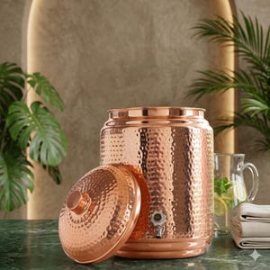 May include: A copper water dispenser with a hammered texture, lid, and tap. The lid is off to the side. The dispenser is on a green surface with plants in the background. A glass jug and folded towels are also visible.