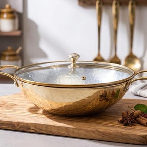Può includere: Una pentola dorata martellata con coperchio in vetro e due manici è appoggiata su un tagliere di legno. Spezie ed erbe sono disposte sulla tavola. Utensili da cucina sono appesi sullo sfondo.