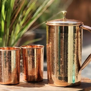 Könnte beinhalten: Ein kupferfarbener Krug mit Deckel und Griff, zusammen mit zwei passenden Bechern. Das Set hat ein geätztes Blumendesign und steht auf einer Holzoberfläche. Der Krug ist etwa 25 cm hoch.