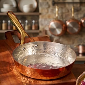 Può includere: Una padella color rame con interno martellato e manico dorato poggia su un tagliere di legno. La padella è rotonda e ha una superficie lucida e riflettente. Utensili da cucina.