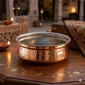May include: A copper-colored bowl with a hammered texture and a silver rim sits on a wooden table. The bowl is round and has a wide, shallow shape. The table has a floral design. The background includes candles and a decorative wooden box.