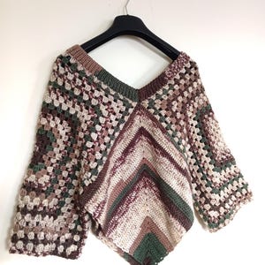 May include: A handmade, crochet poncho in a V-neck style, featuring a mix of earth-tone colours including brown, beige, and green. The poncho is displayed on a black hanger against a white background, showcasing its open weave and flared sleeves.