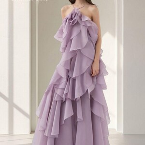 May include: A lavender-colored formal gown with a halter neckline and layered ruffles. The bodice features a large, flower-like embellishment. The dress has a flowing, floor-length skirt. The text "ShamsPoint" is in the upper left corner.