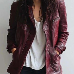 May include: A burgundy leather jacket with a classic design. The jacket features a notched collar, a zipped chest pocket, and button fastenings. The model is wearing a white top and dark blue jeans.