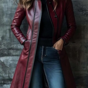 May include: A burgundy leather coat with a longline silhouette. The coat features a mandarin collar, a button closure, and black trim along the seams. The model is wearing a black turtleneck and blue jeans.