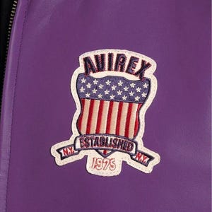 May include: Close-up of a purple leather jacket featuring a patch with the word "AVIREX" above a shield design with red and white stripes and stars. The patch also includes the text "ESTABLISHED 1975" and "N.Y."