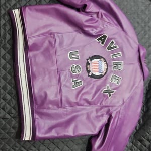 May include: A purple leather jacket with white and black striped cuffs and collar. The back of the jacket features the text "AVIREX USA" with a circular emblem. The jacket is made of a shiny material.