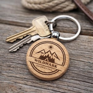 May include: A round wooden keyring featuring a laser-engraved design of a sun, mountain peaks, and a forest. The text WILDMARK ENGRAVING is centred within the circular border. The keyring is attached to a silver metal split ring with two keys.