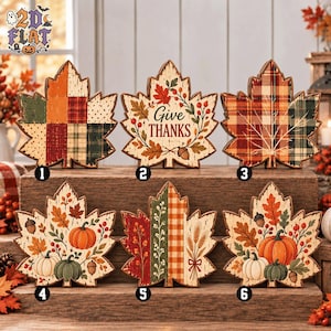May include: Six maple leaf-shaped wooden signs featuring autumn-themed designs. Designs include patchwork patterns, a 'Give THANKS' text, plaid motifs, and illustrations of pumpkins, acorns, berries, and wheat stalks in orange, green, and cream colors.