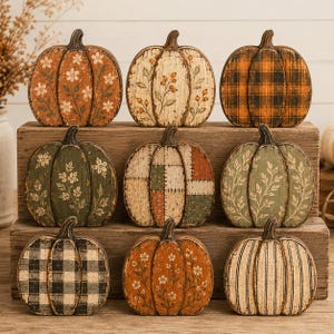May include: A collection of nine decorative wooden pumpkins arranged in three rows. Each pumpkin features distinct patterns including floral prints, plaid, stripes, and patchwork designs in shades of orange, green, black, and cream with a distressed finish.