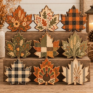 May include: A set of nine decorative wooden leaf-shaped ornaments featuring various autumn-themed designs including floral arrangements, acorns, plaid patterns, a turkey illustration, and wheat stalks with a distressed, rustic finish on each piece.