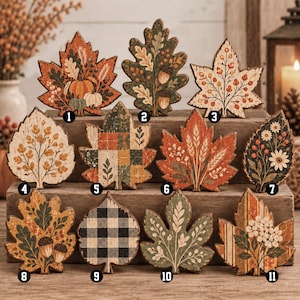 May include: A collection of eleven decorative wooden leaf cutouts featuring various autumn-themed designs, including pumpkins, acorns, wheat stalks, plaid patterns, buffalo check, and floral motifs in shades of orange, green, brown, and cream.