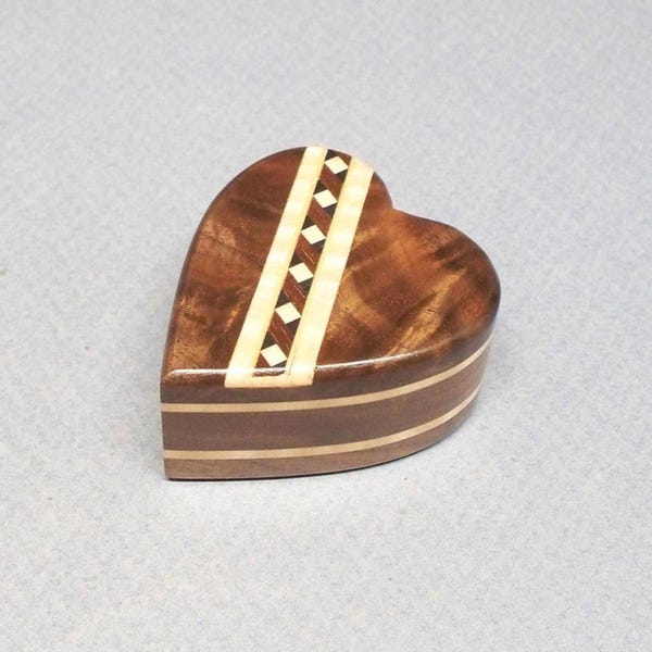 May include: Heart-shaped wooden box with a decorative inlaid design. The box is made of dark brown wood with a lighter wood accent and a geometric pattern. The box is closed and sits on a light gray surface.