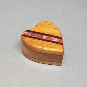 May include: A heart-shaped wooden box featuring a smooth, polished finish with a prominent horizontal inlay of dark wood and a central band of translucent red resin with swirling white patterns, showcasing natural wood grain and layered construction.