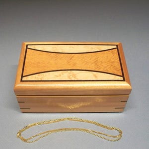 May include: A rectangular wooden jewelry box with a light brown finish and a decorative top. The top features a lighter wood inlay with a curved design, outlined in black. A delicate gold chain necklace rests in front of the box.