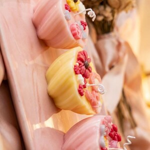 May include: Three decorative candles in the shape of cupcakes, each adorned with colorful floral designs. The candles are pink and yellow, with white wicks and are arranged on a pink surface. A bouquet of dried flowers is in the background.