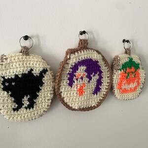May include: Three handmade crochet Halloween ornaments. The ornaments feature a black cat, a purple ghost, and an orange pumpkin. Each ornament has a cream-colored center with a brown border and a metal ring for hanging.