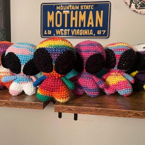 May include: A collection of handmade crochet alien dolls in various colors, including rainbow, pink, and purple. Each doll has large black ears and a small body. The dolls are displayed on a wooden shelf, with a Mothman license plate in the background.