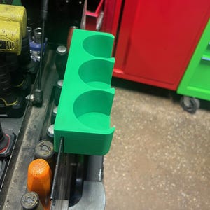 May include: A bright green, rectangular tool organizer featuring three circular cutouts designed to hold cylindrical items. The organizer is mounted onto the side of a dark metal tool cart, positioned vertically with a flat base and open top.