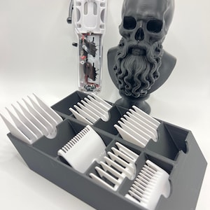 May include: A clear and white hair clipper with the text "TPOB" and a black skull bust with a long beard. A gray organizer holds several white clipper guards. The clipper guards are of various sizes.