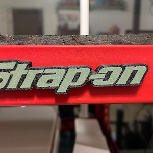 Op de afbeelding: Een felrood metalen oppervlak met een verhoogd, driedimensionaal logo met de tekst "Strap-on" in een gestileerd lettertype. De letters hebben een getextureerde, licht saliegroene voorzijde met een dikke zwarte rand, horizontaal gemonteerd.