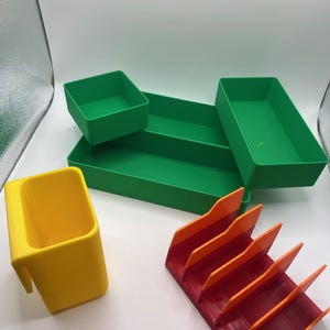 Puede incluir: Un surtido de organizadores impresos en 3D en varios colores. Incluye una bandeja rectangular verde con dos compartimentos, un pequeño recipiente cuadrado verde, una taza amarilla con asa y un organizador angular rojo y naranja.