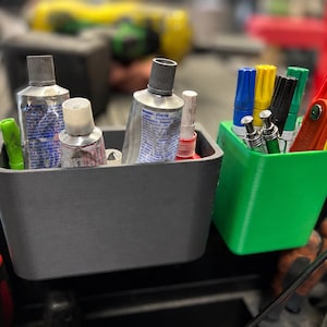 Op de afbeelding: Twee plastic opbergcontainers, één grijs en één groen, bevatten diverse gereedschappen en benodigdheden. De grijze container bevat lijmtubes en een groene marker. De groene container bevat markers, inbussleutels en ander gereedschap.