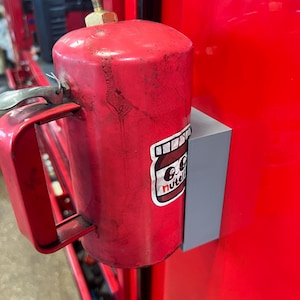 May include: A red cylindrical metal canister with a handle and a brass-colored top fitting, mounted to a red surface by a gray rectangular bracket. A sticker featuring a cartoon jar with the text Nutella is applied to the side of the canister.