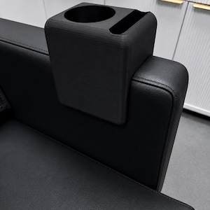 May include: A matte black couch armrest organizer featuring a circular cup holder and a rectangular slot for remote controls or mobile devices. The organizer is constructed with a saddle-style design that fits securely over the arm of a black sofa.