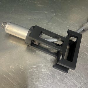 Może przedstawiać: Czarny i srebrny element mechaniczny ze sprężyną, prostokątną ramą i metalowym prętem. Część ma wydrukowany numer "1267029". Obiekt znajduje się na metalowej powierzchni.