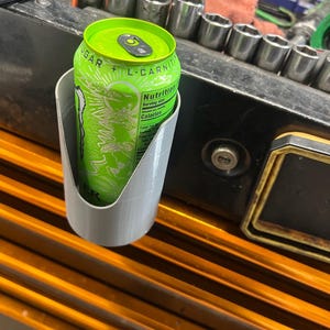 May include: A light gray cylindrical cup holder with a U-shaped cutout on the front, mounted to a surface. A bright green energy drink can with the text "SUGAR + L-CARNITINE" and "Nutrition" printed on the label is placed inside the holder.