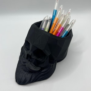 May include: A matte black, geometric, low-poly skull-shaped desk organizer. The hollow top section holds a collection of various pens with clear plastic barrels and colorful ink caps, including blue, orange, yellow, pink, and light blue accents.