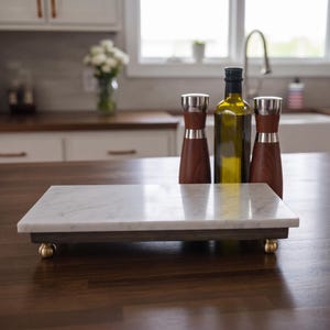 May include: A rectangular white marble serving board with grey veining, featuring a dark wood base and four small brass-toned ball feet. Behind the board stand two wooden salt and pepper grinders with silver accents and a glass bottle of olive oil.