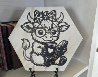Può includere: Una piastrella decorativa esagonale in bianco e nero con una mucca dei cartoni animati con occhiali e un fiocco, che legge un libro con un cuore sulla copertina. La piastrella è esposta su un supporto in metallo nero.