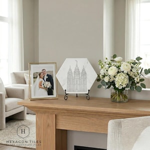 May include: A gold framed photo of a couple in wedding attire sits next to a white hexagonal tile with a laser-engraved architectural design. The tile is displayed on a black metal stand. A vase of white flowers is also on the wooden surface.