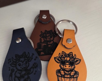 Può includere: Tre portachiavi in pelle blu navy, marrone e arancione. Ogni portachiavi presenta un'illustrazione di mucca. Il portachiavi blu navy mostra una mucca che canta con un microfono. Il portachiavi marrone mostra una mucca che guida un trattore. Il portachiavi arancione mostra una mucca in una botte con occhiali da sole a forma di cuore.