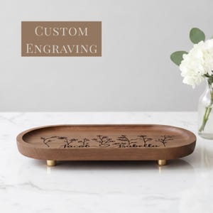 May include: An oval wooden tray with a smooth brown finish, featuring a custom engraving of wildflowers and the names Jacob and Isabella. The tray is supported by four small, cylindrical gold-toned metal feet on the underside.