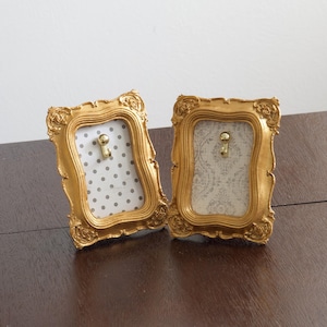 May include: Two gold-colored ornate frames with decorative scrollwork edges. Each frame features a central gold hook mounted on a backing. One frame contains a white background with gray polka dots, while the other has a white and gray damask pattern.