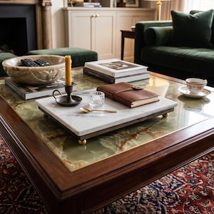 May include: A rectangular white marble serving tray with a dark wood base and four small brass ball feet. The tray holds a dark metal candle holder with a yellow taper candle, a small clear glass votive, a gold spoon, and a brown leather journal.