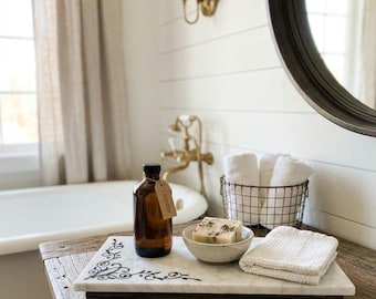Può includere: Un vassoio rettangolare in marmo bianco con angoli a volute nere e piedini in ottone, che sostiene una bottiglia in vetro ambrato con etichetta alla lavanda, una piccola ciotola in ceramica con una saponetta e un panno bianco piegato.