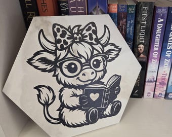 Può includere: Cartello in legno esagonale con una mucca dei cartoni animati in bianco e nero con occhiali e un fiocco, che legge un libro con un cuore. Il cartello è davanti a una libreria piena di libri.
