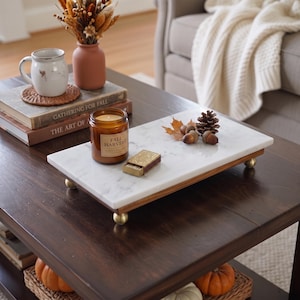 May include: A rectangular white marble serving tray with a natural wood base and four small brass ball feet. The tray holds an amber glass candle jar labeled Fall Harvest, a small rectangular matchbox, a pinecone, and two dried maple leaves.